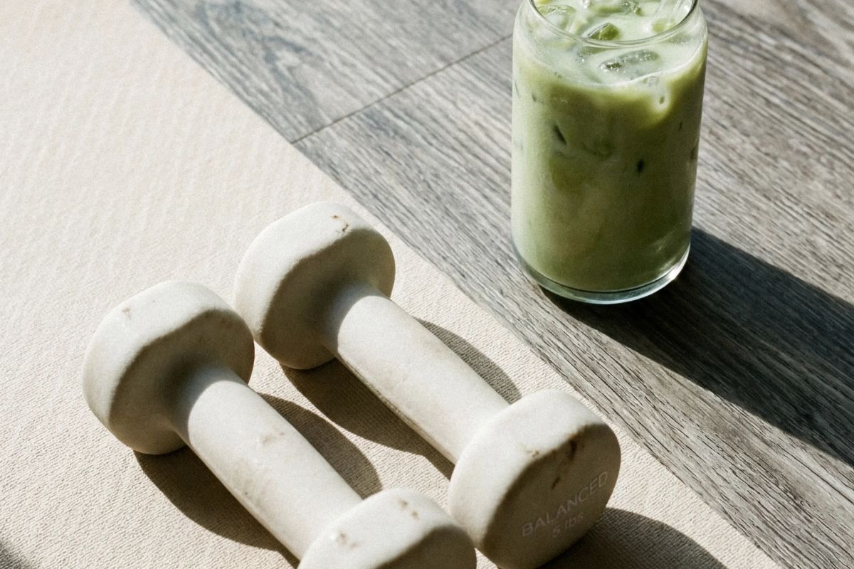 Cream dumbbells resting on a cream yoga mat beside an iced matcha drink on a light gray wood floor