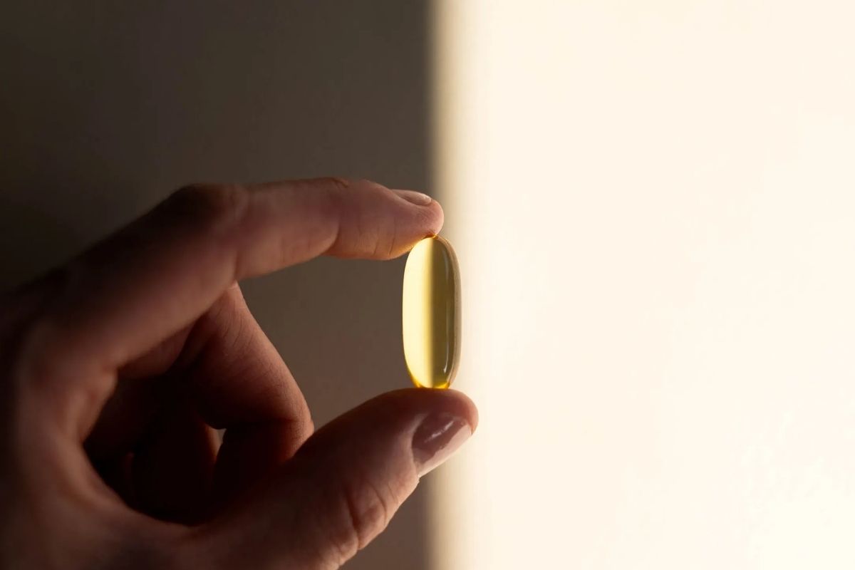 A hand holding a single golden softgel capsule upright with warm sunlit light glowing behind it