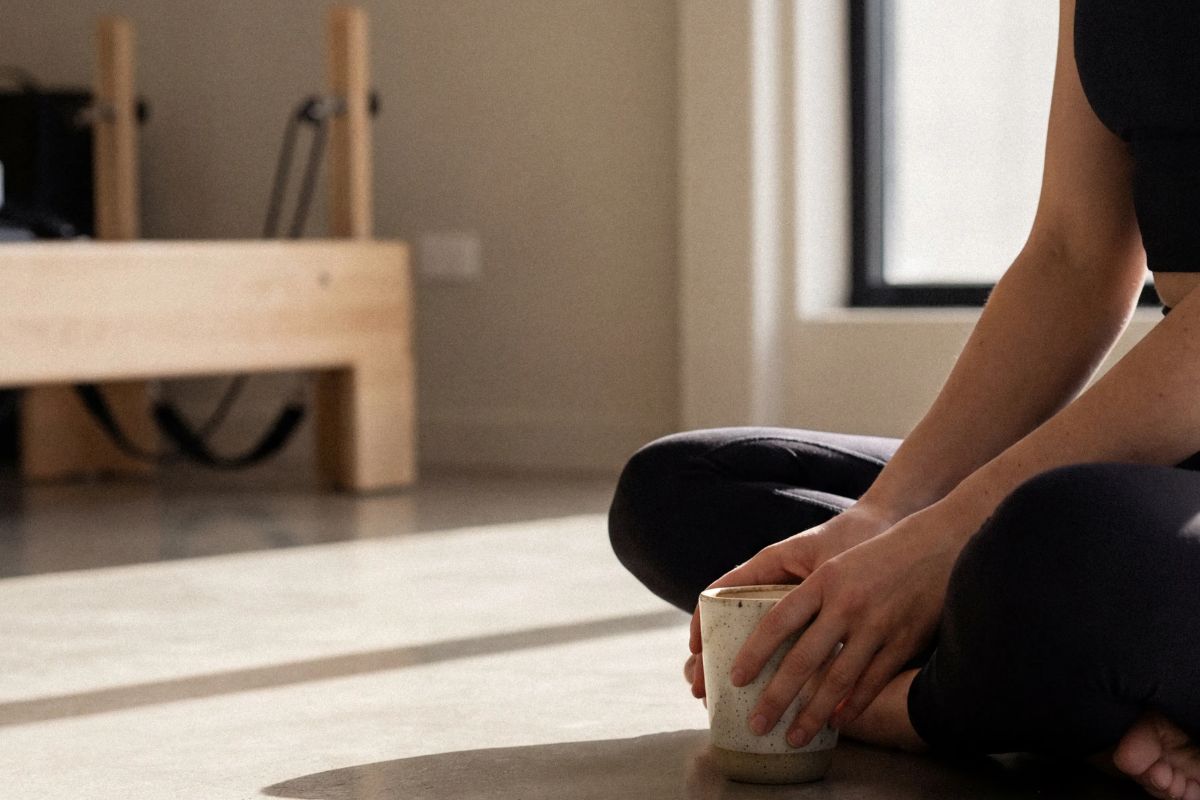 A woman seated in a Pilates studio cradling a cream ceramic mug with a reformer and light wood floor visible beside a sunlit window