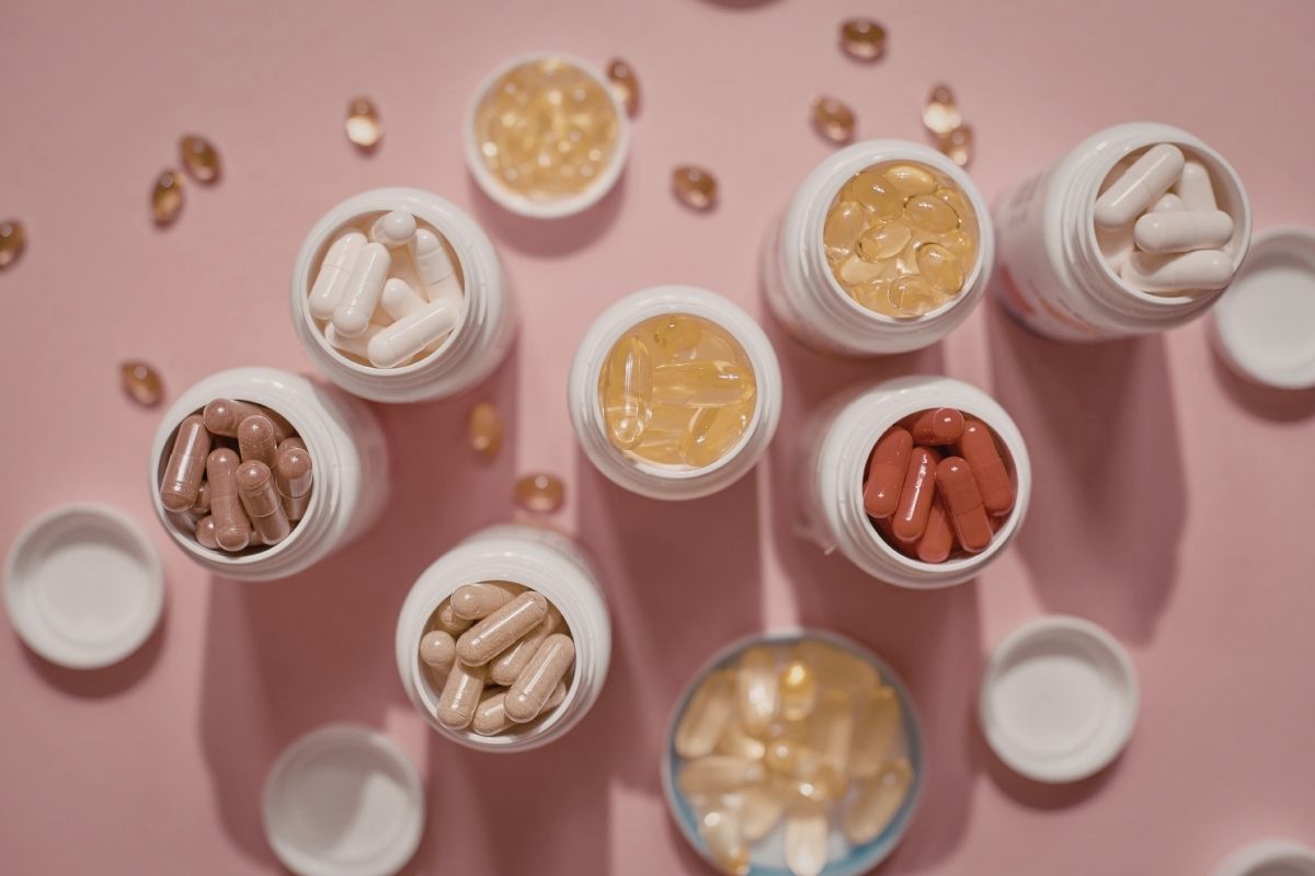 A curated collection of supplement bottles and capsules in various colors and sizes arranged on a soft pink background.