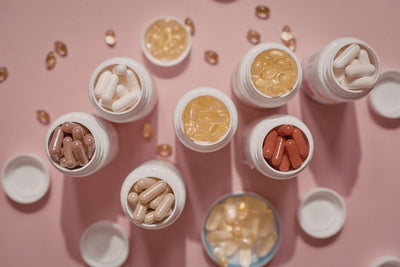 A curated collection of supplement bottles and capsules in various colors and sizes arranged on a soft pink background.